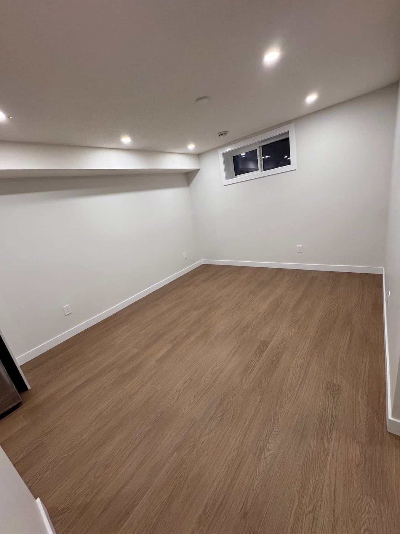 The same basement living area at handover: vinyl plank flooring down, floor and walls cleaned, room empty and ready for occupancy.