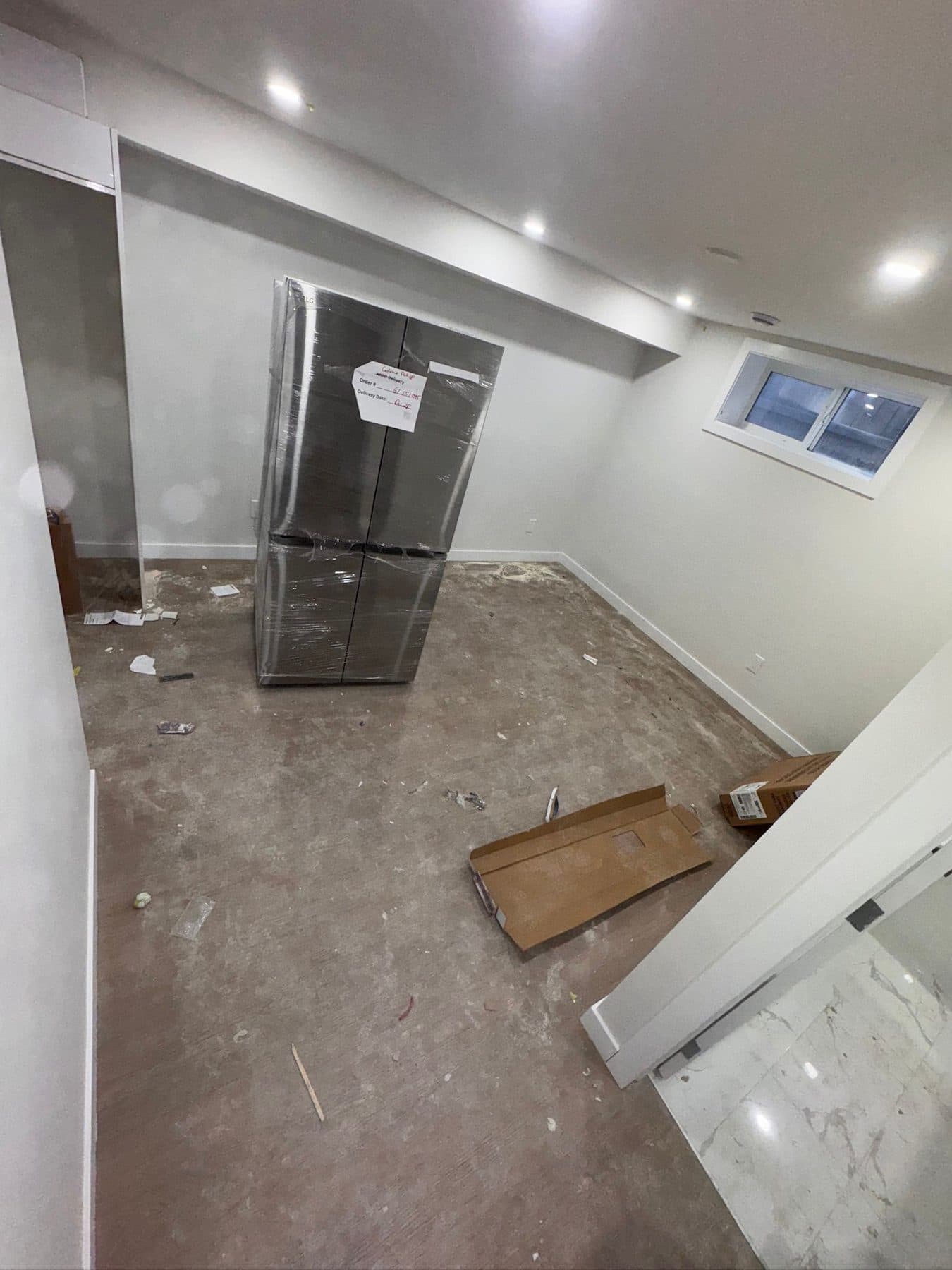 Basement living area at site condition: bare concrete floor with construction debris, offcuts and dust, appliance still standing in the room.